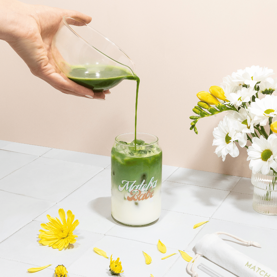 Hand pouring green matcha through the glass bowl’s spout into a Matcha Babe glass of iced milk, beside white and yellow flowers.