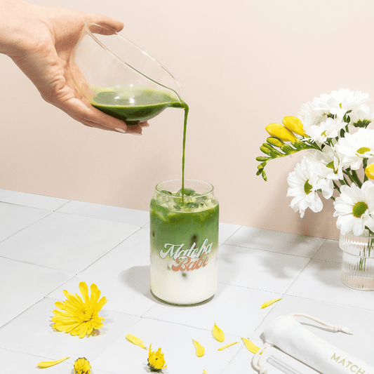 Hand pouring green matcha through the glass bowl’s spout into a Matcha Babe glass of iced milk, beside white and yellow flowers.