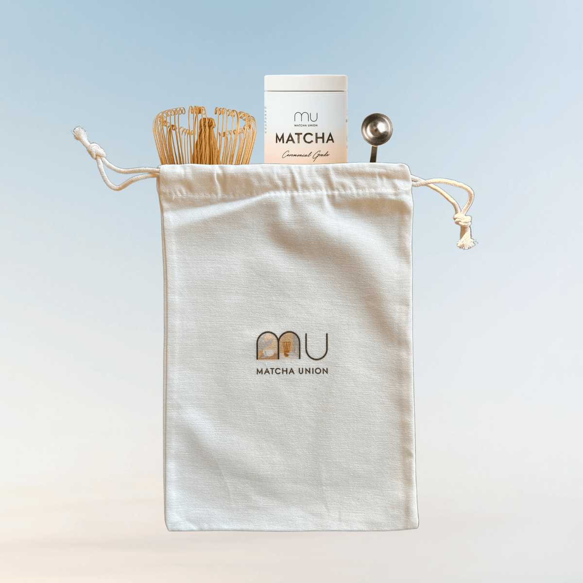 Matcha Union cotton drawstring gift bag holding a ceremonial matcha tin, bamboo whisk and metal scoop against a pale blue background.