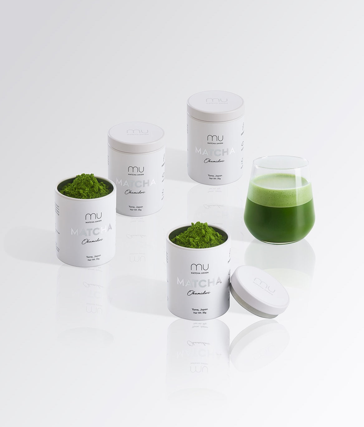 Four Matcha Union Okumidori tins, two open with green matcha powder, beside a glass of prepared matcha.