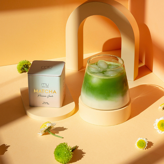 Matcha Union Premium Matcha tin beside a layered iced matcha latte on peach display stands, surrounded by flowers in warm sunlight.