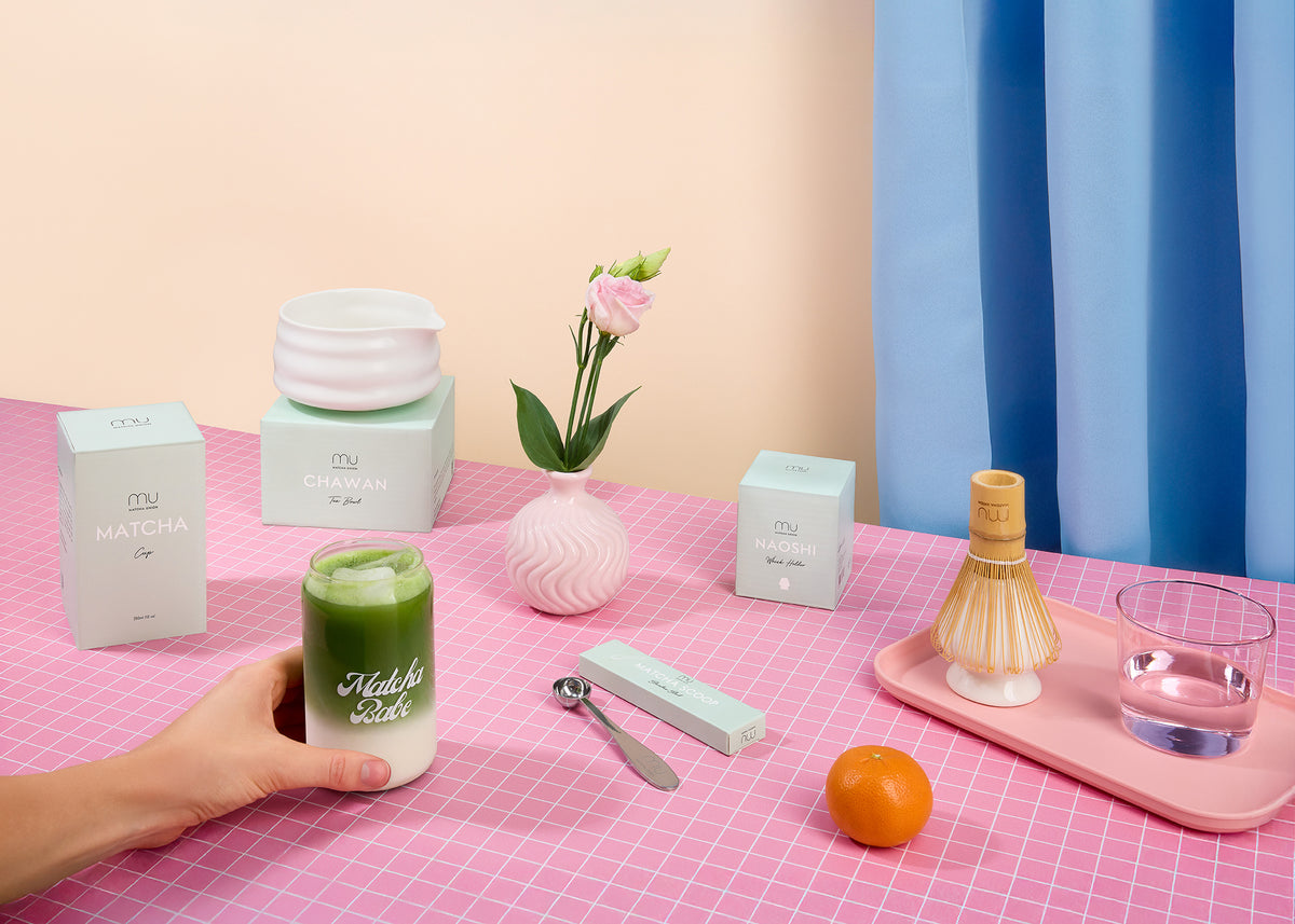 Hand holding a Matcha Babe glass of iced matcha latte on a pink table, with a ceramic bowl, bamboo whisk, scoop and Matcha Union packaging nearby.