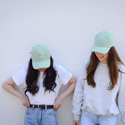 Two people wearing matcha-green Energise Focus Glow caps with white embroidered symbols, styled with white tops and blue jeans.