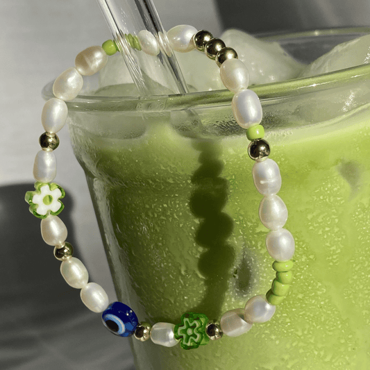 Close-up of the Matcha Magic bracelet draped over an iced matcha glass, showing freshwater pearls, gold beads, green flowers and a blue eye bead.
