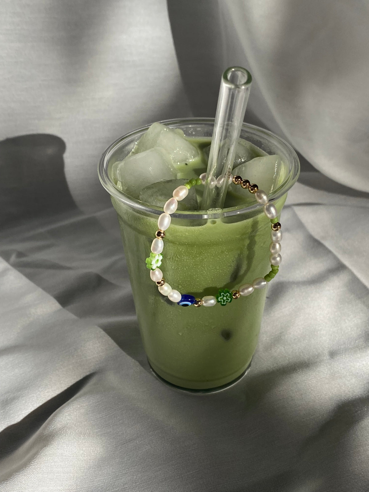 Matcha Magic pearl bracelet hanging over the rim of a tall iced matcha latte, with green flower beads and a blue eye bead facing forward.
