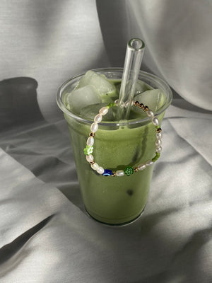 Matcha Magic pearl bracelet hanging over the rim of a tall iced matcha latte, with green flower beads and a blue eye bead facing forward.