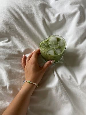 Hand holding a ribbed glass of iced matcha latte while wearing the Matcha Magic bracelet, with pearl, gold and green glass beads visible.