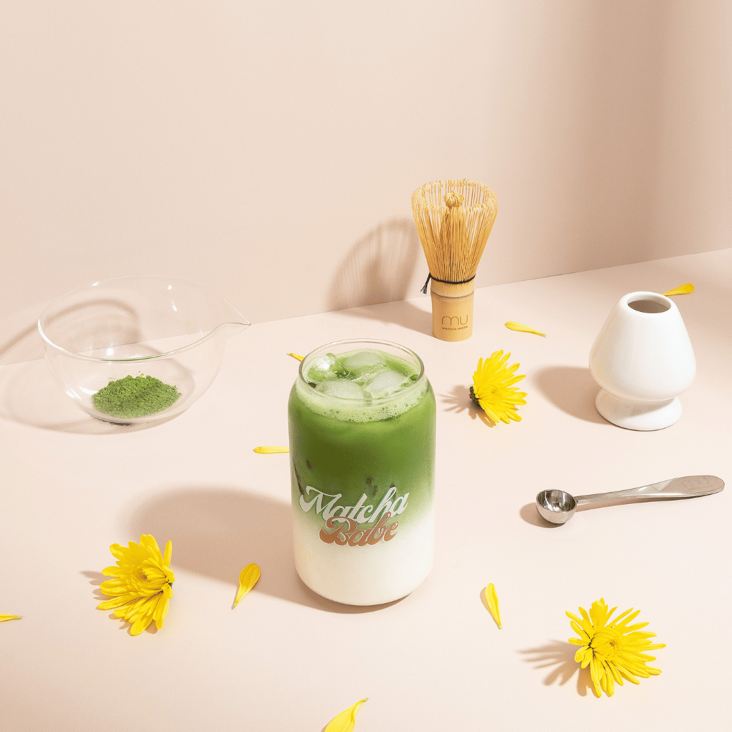 Stainless steel matcha scoop beside an iced matcha latte, bamboo whisk, white whisk holder and glass bowl of matcha powder on a peach surface with yellow flowers.