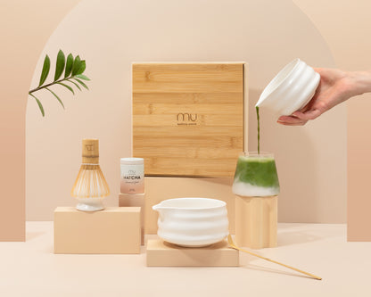 Hand pouring matcha from a white ceramic bowl into a glass of milk, with the Matcha Union Ceremony Set’s matcha tin, whisk, holder, scoop and bamboo-lidded box displayed nearby.