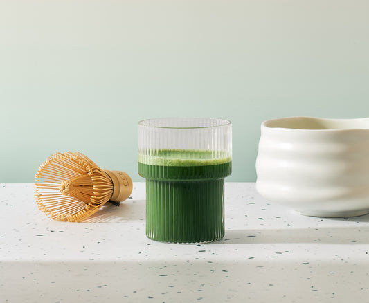 White ceramic Japanese tea bowl beside a ribbed glass of green matcha and a bamboo whisk on a speckled countertop.
