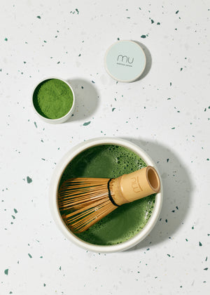 Overhead view of frothy green matcha in a white ceramic chawan with a bamboo whisk resting inside, beside an open tin of matcha powder.