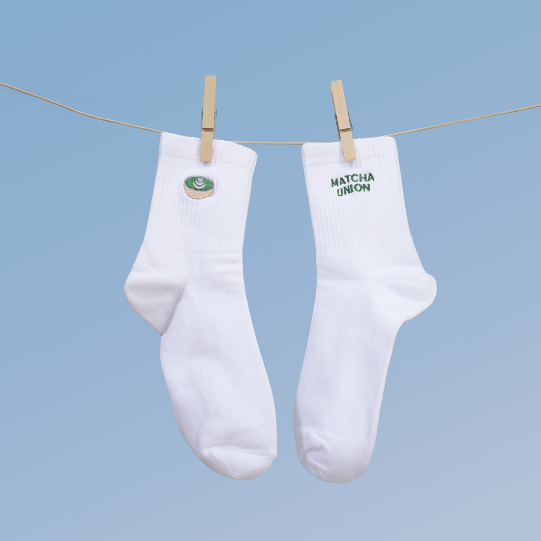 White Matcha Union socks hanging from wooden pegs against a blue background, showing the embroidered matcha cup and green brand lettering.