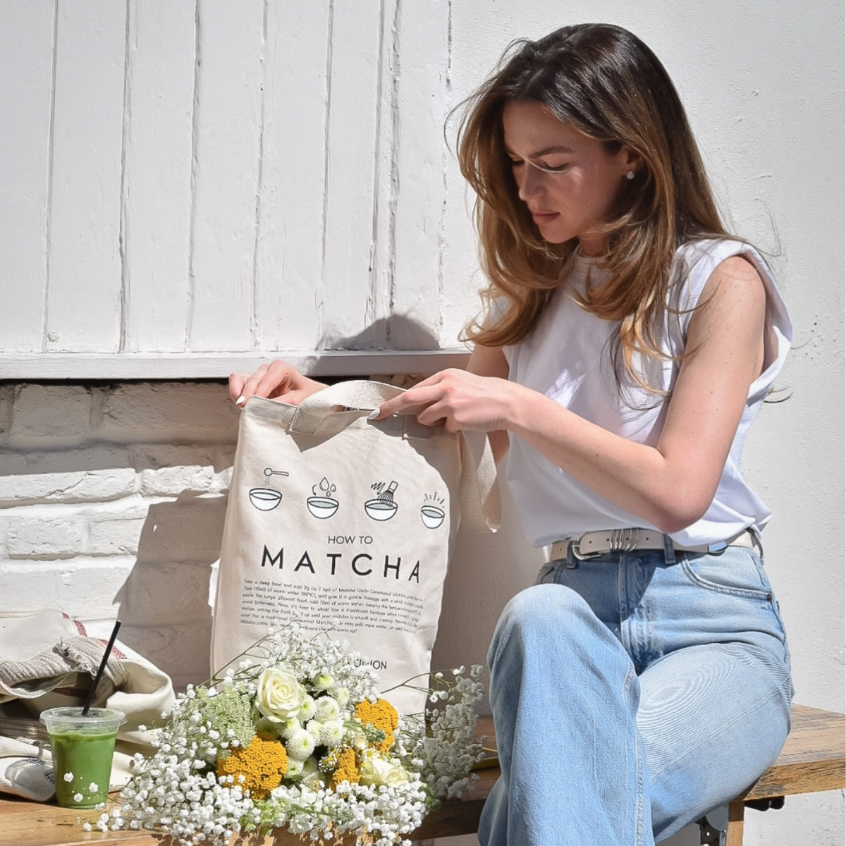 Person seated on a bench holding a roomy Matcha Union tote bag with its matcha preparation illustration visible, beside flowers and a green matcha drink.