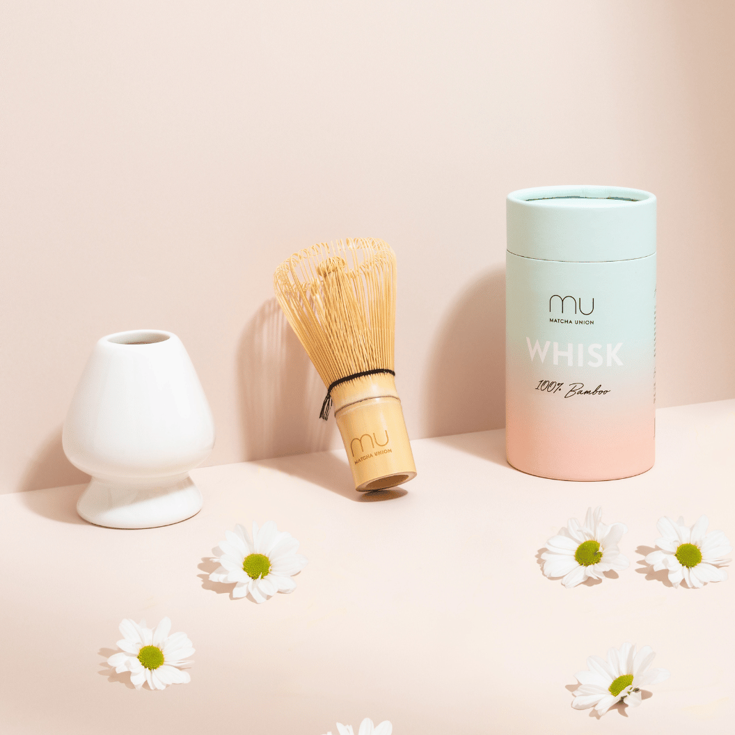Matcha Union Bambusbesen neben einem weißen Keramikhalter und einer pastellfarbenen Besen-Hülle, mit weißen Gänseblümchen auf pfirsichfarbenem Display.