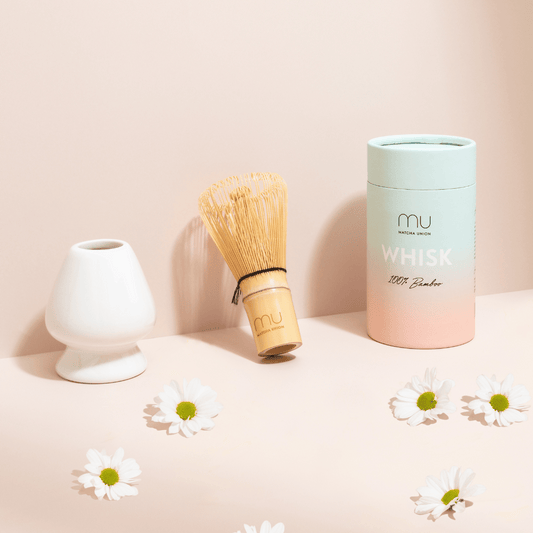 Matcha Union bamboo whisk beside a white ceramic holder and pastel whisk tube, with white daisies on a peach-coloured display.