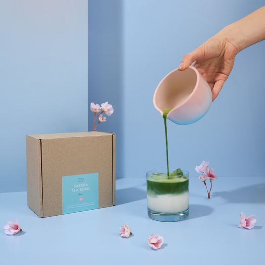 Hand pouring green matcha from the Sakura stoneware bowl into a glass of iced milk, beside its box and pink blossoms on a blue display.