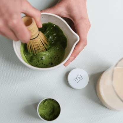 Blick von oben auf Hände, die mit einem Bambusbesen schaumigen grünen Matcha in einer weißen Schale mit Ausgießer aufschlagen, daneben eine offene Matcha-Dose und ein Glas Milch.