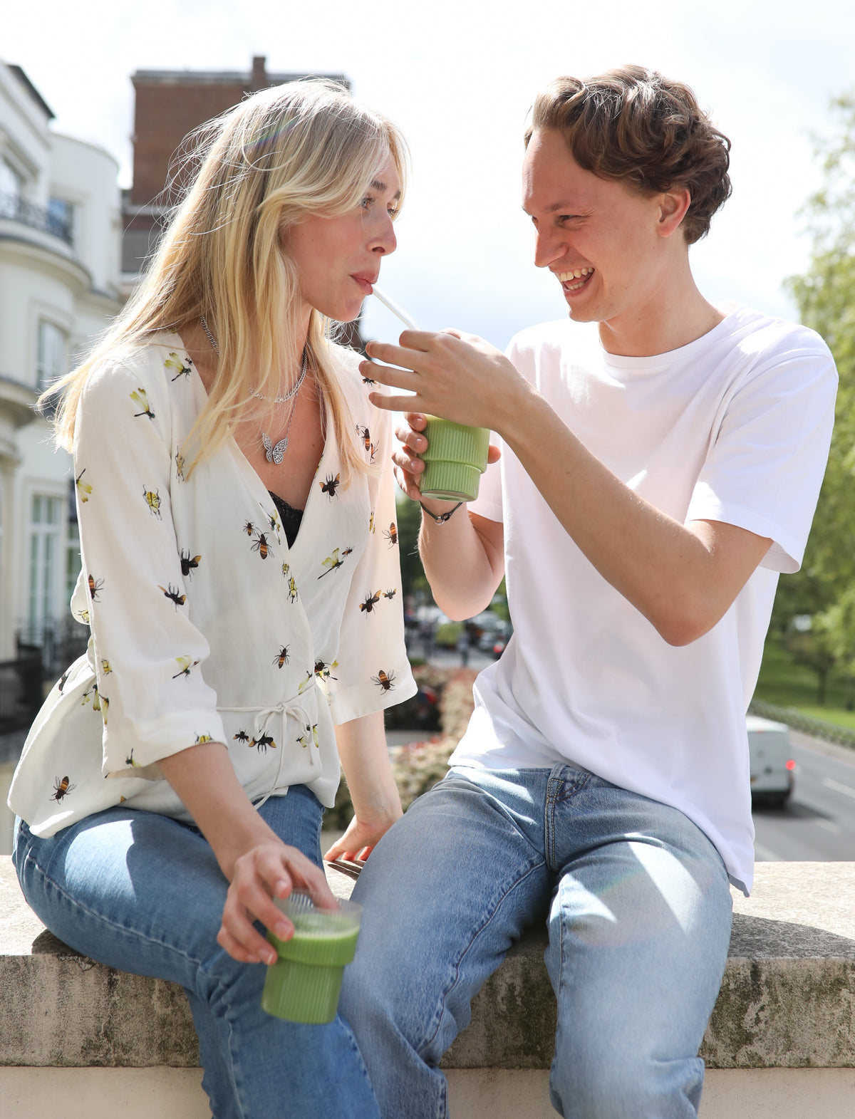 Two people sitting on a brick wall outdoors, smiling and holding ribbed glasses of green matcha.