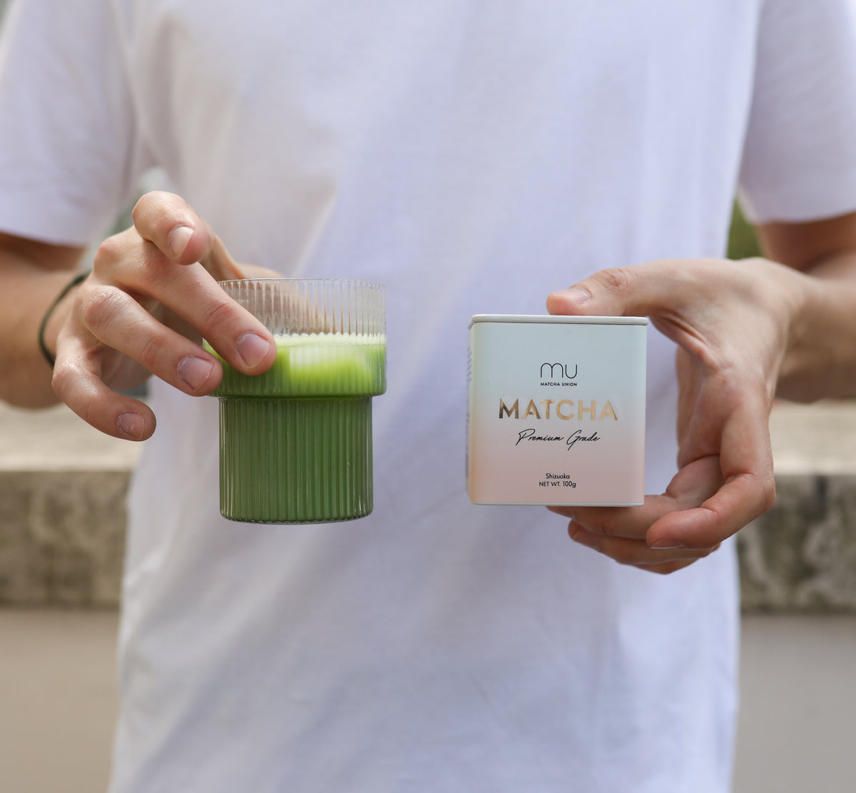 Person holding a Matcha Union Premium Matcha 100g tin alongside a ribbed glass of green matcha topped with a light layer of foam.